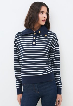 Vrouw draagt een navy-wit gestreept shirt met lange mouwen, een brede navy kraag en gouden knopen, gecombineerd met donkerblauwe jeans, kijkt naar rechts.