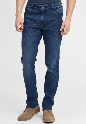Mørkeblå denimbukser med rett passform, fem-lomme-design, lett slitasje og knappelukking. Kombinert med lysebrune sko.