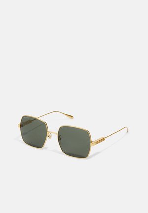 Gafas de sol de metal dorado con lentes cuadrados, cristales verdes y un detalle texturizado en el costado que presenta el nombre de la marca en letras en 3D.