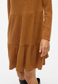 Robe en tricot côtelé marron avec des manches longues, un ourlet évasé et un design à étagères. La texture est douce ; la couleur est chaude et terreuse.