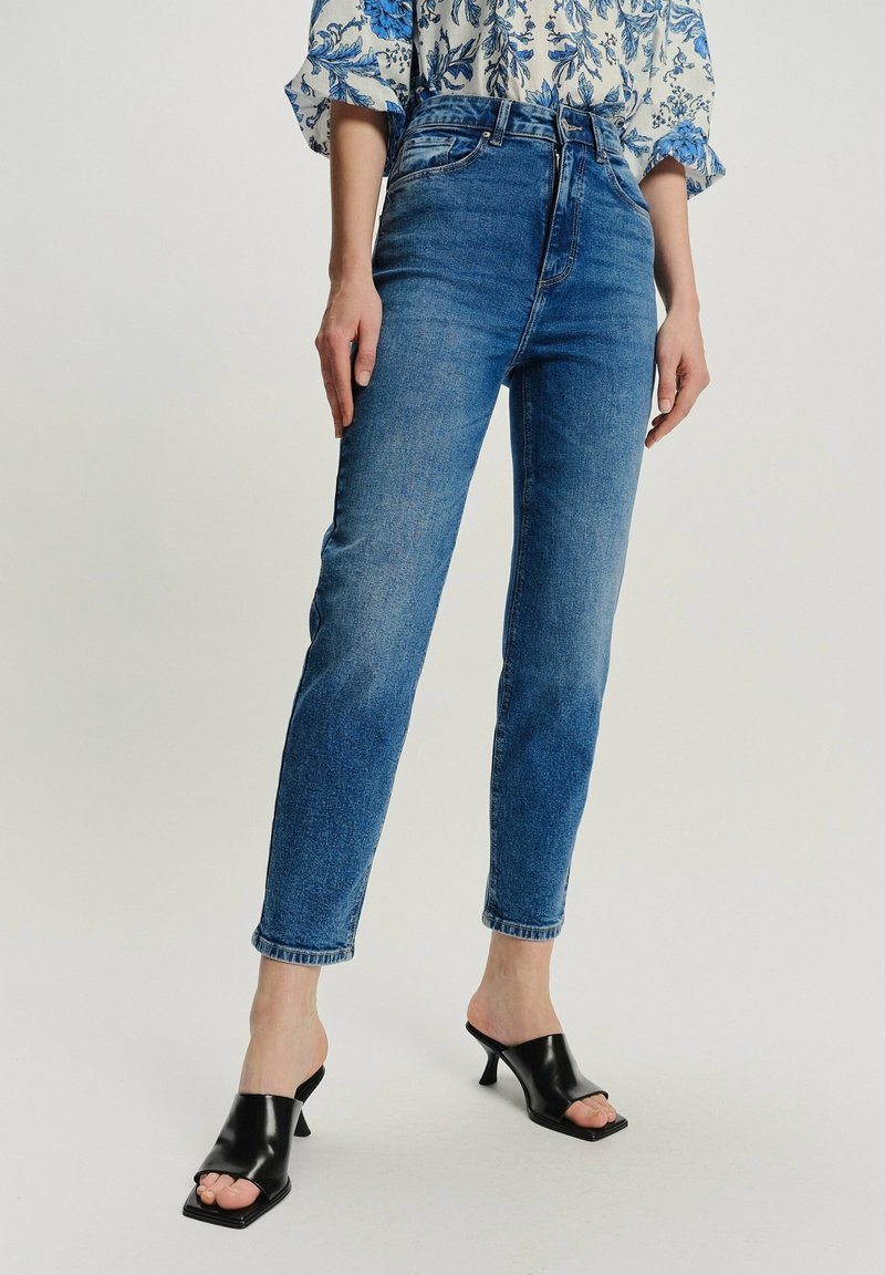Mujer con jeans ajustados de tiro alto azul, mule de tacón negro y blusa floral azul y blanca, de pie con las manos relajadas a los lados.