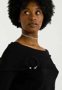 Mujer con cabello rizado que lleva una blusa negra de hombros al descubierto con un detalle metálico circular y un collar de strass, mirando de lado.