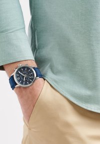 Orologio cronografo argento con cinturino in silicone blu, quadrante blu scuro, indicatori delle ore bianchi e dettagli accentuati; indossato con una camicia verde menta.