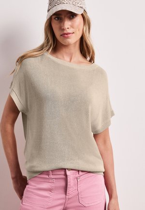 Donna con lunghi capelli biondi che indossa una maglia beige a maniche corte lavorata a maglia, jeans rosa e un berretto chiaro con stampa animalier, in piedi davanti a uno sfondo uniforme.