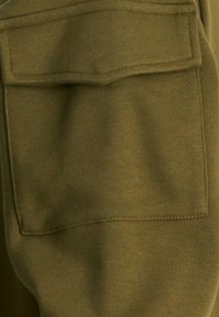 Tejido de sudadera verde oliva con una textura acanalada, que presenta un bolsillo de parche cuadrado cosido en la parte posterior. La costura es visible a lo largo de los bordes.
