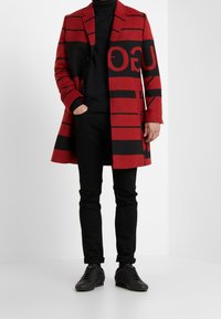 Czerwono-czarny wełniany płaszcz z odważnymi poziomymi paskami, jednorzędowy fason, dopasowany krój, zestawiony z czarnymi spodniami i butami.