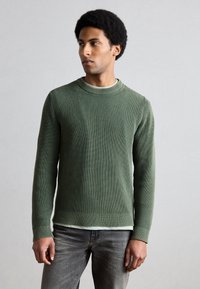 Zeleno rebrast pulover z okroglim izrezom in dolgimi rokavi, nošen čez svetlo barvno srajco, kombiniran s sivimi kavbojkami.