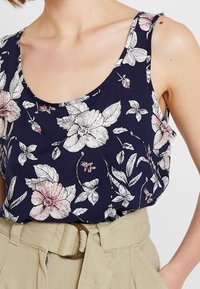 Ärmeloses Blumenshirt aus marineblauem Stoff, mit großen rosa und weißen Blumen sowie grünen Blättern, Rundhalsausschnitt und weicher Textur.
