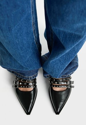 Chaussures noires à bout pointu avec découpes, dotées de quincaillerie métallique et de sangles cloutées, associées à un jean en denim bleu légèrement évasé.