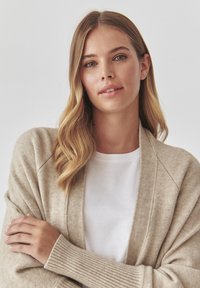 Cardigan beige avec des poignets côtelés, à coupe décontractée et à l'avant ouvert, superposé sur un t-shirt blanc. Tissu doux et texturé.