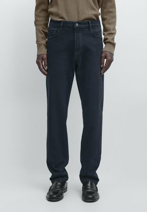 Person wearing dark blue straight-leg jeans, brown ribbed sweater, and black leather loafers, standing against a plain light background.