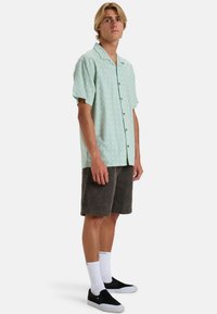 Maglietta a maniche corte di colore verde menta con un sottile motivo a foglia, design con bottoni, abbinata a pantaloni corti in velluto a coste grigi e scarpe nere slip-on.