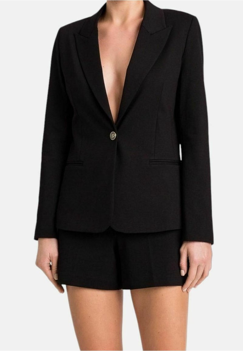 Femme portant un blazer noir cintré avec un seul bouton et un short noir assorti, debout devant un fond uni.