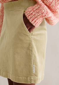 Lichtgroene corduroy rok met zijzakken, getextureerde ribbels en een klein merkembleem bij de zoom. Gestyled met een roze, gebreide trui.