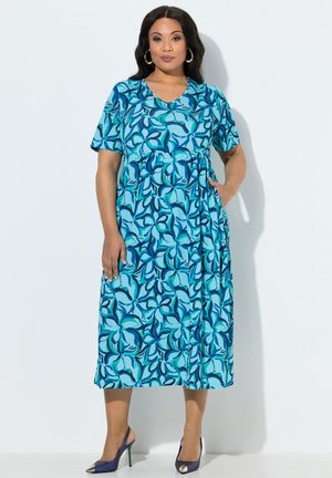 Frau trägt ein blaues Midikleid mit Blumenmuster und kurzen Ärmeln, eine Hand in der Tasche, dazu dunkle High-Heels vor weißem Hintergrund.