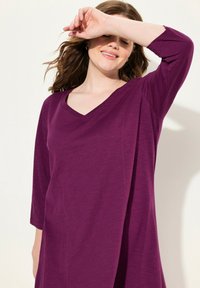 V-kaulus violetinvärinen mekko, jossa on kolmannen osan hihat, pehmeä rakenne, virtaava siluetti ja vähäiset ompeluyksityiskohdat.