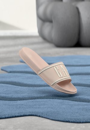 Sandale à glissière beige avec des lettres blanches en relief "GAP" sur la lanière, posée sur un tapis bleu texturé, fond gris doux.