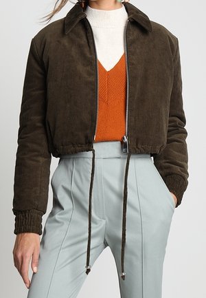 Giacca cropped in corduroy marrone con zip frontale, orlo raccolto e colletto. Indossata sopra un maglione arancione con texture e pantaloni azzurri chiari.
