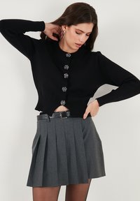 Black knitted cardigan with a round neckline, featuring six ornate silver buttons. Paired with a pleated gray skirt and a black belt.