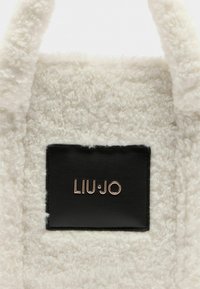 Borsa color crema con texture in peluche, caratterizzata da un'etichetta in pelle nera con la scritta "LIU·JO" in oro metallico. Presenta manici rotondi.