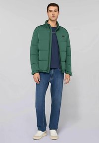 Grön vadderad jacka med hög krage, hel dragkedja och logotypmärke. Bärs över en marinblå skjorta och kombineras med blå jeans och vita sneakers.