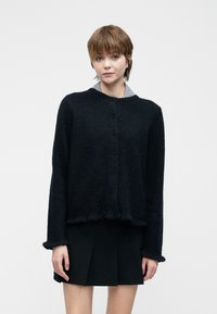 Žena s krátkými vlasy, oblečená v černém propínacím svetru přes plisovanou černou sukni, stojí proti jednoduchému bílému pozadí.