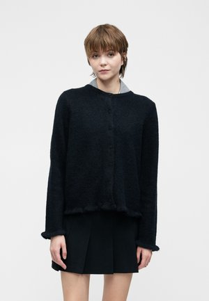 Femme aux cheveux courts portant un cardigan noir à boutons sur une jupe plissée noire, se tenant devant un fond blanc uni.