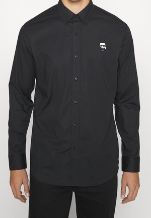 Mężczyzna ubrany w czarną koszulę na guziki z małym, białym i czarnym stylizowanym logo twarzy na lewej piersi.