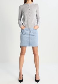 Ljusgrå långärmad cardigan med knäppning och ribbade muddar, kombinerad med en blå jeanskjol med fransad kant. Svarta högklackade skor.