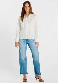 Wit katoenen overhemd met knopen en kraag, gecombineerd met lichtblauwe wijde jeans. Model staat voor een neutrale achtergrond en draagt bruine sandalen met hak.