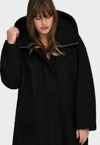 Črn plašč iz volnenega mešanega materiala z veliko kapuco, teksturirano površino in zadrgo po celotni dolžini. Ima spuščena ramena in sproščen kroj.