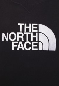 Tissu noir avec texte brodé blanc "THE NORTH FACE" et trois lignes courbées formant un quart de cercle vers la droite.