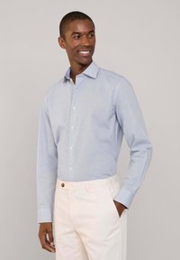 Camisa de botones azul clara con un pequeño patrón de cuadros, mangas largas y cuello, combinada con pantalones color crema que tienen un botón marrón.