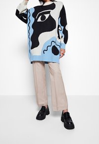 Överdimensionerad stickad tröja i kräm, blå, svart och vit med abstrakt öga-design. Matchad med ljusbeige vida byxor och klumpiga svarta loafers.