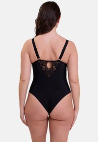 Femme aux cheveux longs portant un body en dentelle noire, vue de dos contre un fond blanc uni.