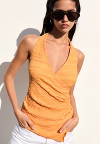 Oranžový halter top s texturou a hlubokým V-krkem a sklady. V kombinaci s bílými šortkami a elegantními černými slunečními brýlemi.