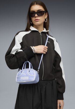 Femme portant une veste Puma noire et blanche, des lunettes de soleil noires, des boucles d'oreilles argentées en forme d'anneau, des bracelets, et tenant un petit sac à main Puma violet clair.