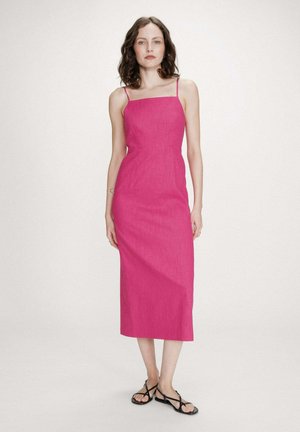 Femme portant une robe midi rose vif à fines bretelles et des sandales plates noires à brides, debout devant un fond clair uni.