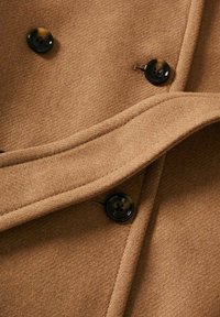 Tissu de manteau marron avec un gros plan sur des boutons de style écaille de tortue et des coutures détaillées le long du col et des coutures.