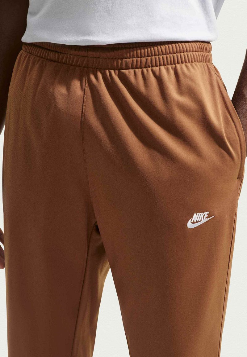 Brune Nike joggingbukser med elastisk talje og sidelommer, båret med hvid top, der viser Nike-logoet på højre lår.