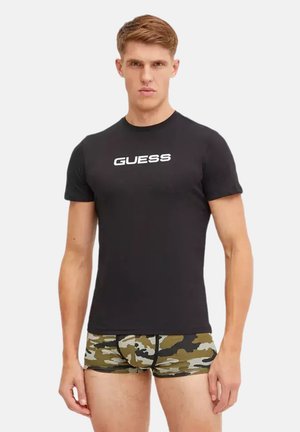 Jeune homme portant un t-shirt noir ajusté de la marque Guess et un boxer camouflage vert, marron et noir, debout devant un fond uni.