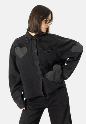 Sort denim oversized skjorte med hjerteformede lapper på brystet og ærmerne, sættet med sorte denim bukser. Har en krave-udskæring og knaplukning.