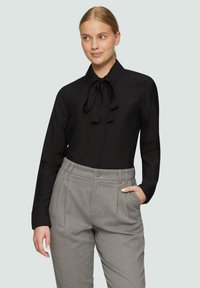 Schwarze Bluse mit langen Ärmeln und einer gebundenen Schleife am Kragen, kombiniert mit grauen, hochgeschnittenen Hose mit Falten und Seitentaschen.
