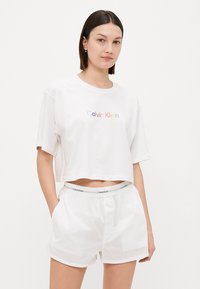 Hvit crop top med et flerfarget "Calvin Klein"-logo. Kombinert med hvite shorts, elastisk midje, glatt tekstur og uformell design.