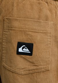 Ruskea mantereinen vetoketjutasku, jossa on mustavalkoinen Quiksilverin logopäällinen ommeltuna kangaskappaleeseen.