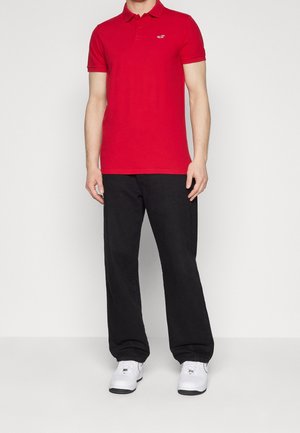 Polo rouge à manches courtes, texture lisse, avec un logo discret sur la poitrine gauche, associé à un pantalon ample noir et des baskets blanches.