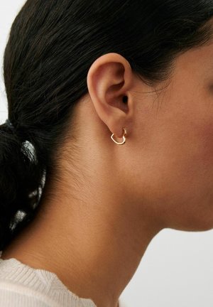 Lähivõte tumedate juustega naise kõrvast, juuksed tagant seotud, kannab väikest ebaühtlaselt kujundatud kuldset rõngaskõrvarõngast.