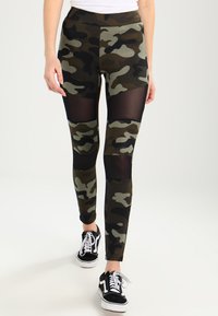 Kamouflageleggings i grönt och svart med nätpaneler, hög midja och åtsittande design. Kombinerat med svarta sneakers med vita snören.