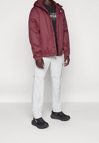 Bordowa lekka kurtka z kapturem, czarny t-shirt z grafiką pod spodem, białe spodnie z zamkiem błyskawicznym oraz czarne buty sportowe z żółtymi akcentami.