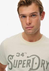 Camiseta de color crema claro con un gran estampado gris de estilo vintage. Diseño de cuello redondo y mangas cortas, con una textura de tela suave.